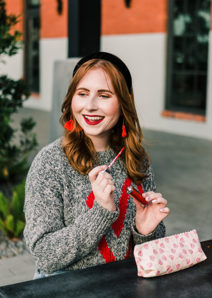 Valentine's Day Drugstore Makeup Look. Amanda Burrows of Affordable by Amanda is wearing a heart gray sweater from LOFT. She is applying red lipstick from Milani. 