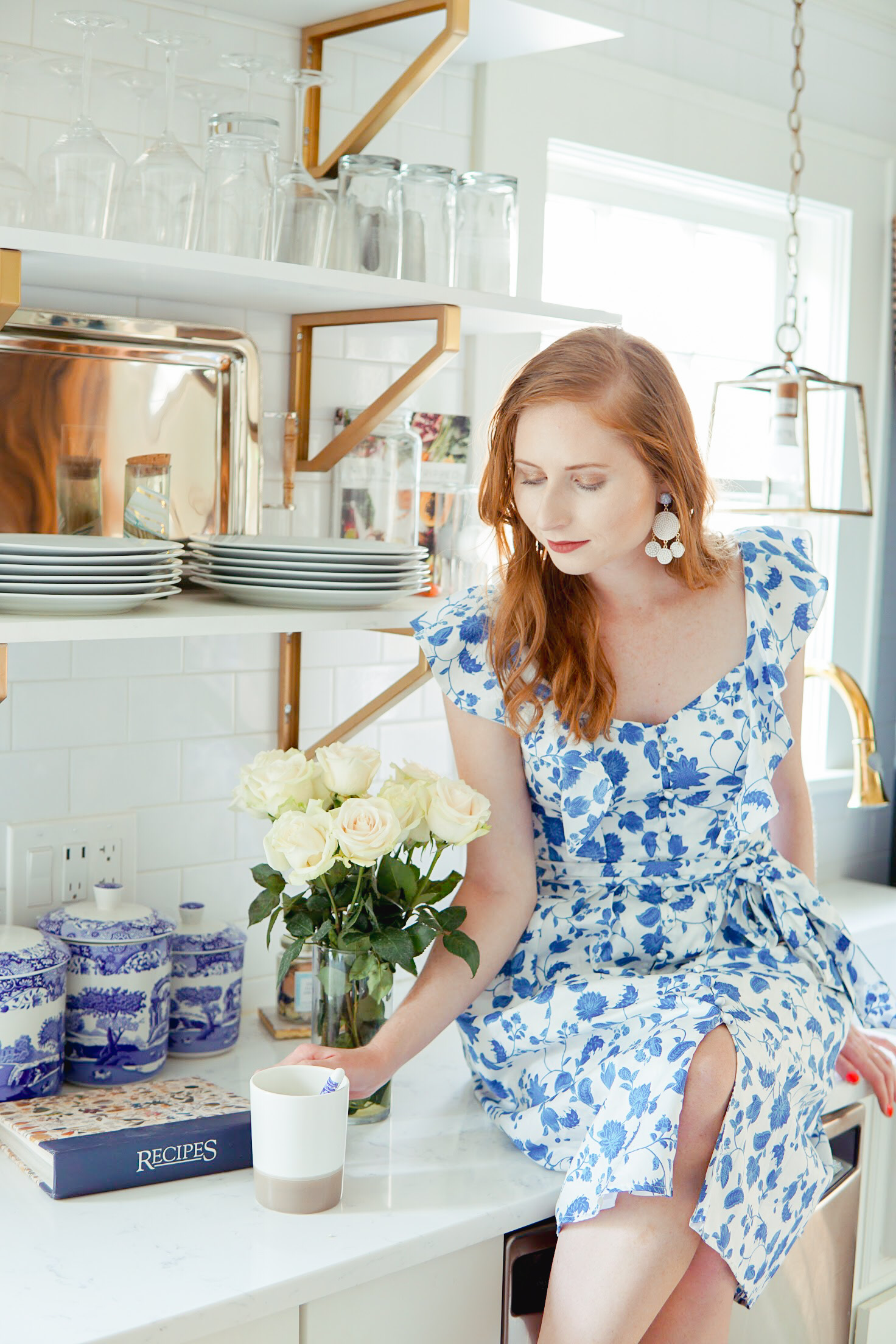 Tips for Working From Home | Amanda Burrows holds a cup of tea inside kitchen. She is wearing a blue and white Gal Meets Glam dress.