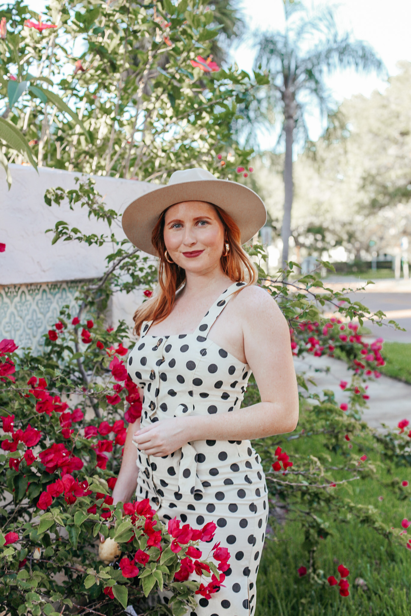 A Budget-Friendly Everyday Dress. Florida style blogger Amanda Burrows of the fashion blog Affordable by Amanda shares her favorite Spring Dresses Under $50. Amanda wears a casual JustFab Belted Button Front Dress with polka dot print and an Urban Outfitters Wide Brim Beige Hat. She is standing in a bush of bougainvillea in St. Petersburg, Florida. 