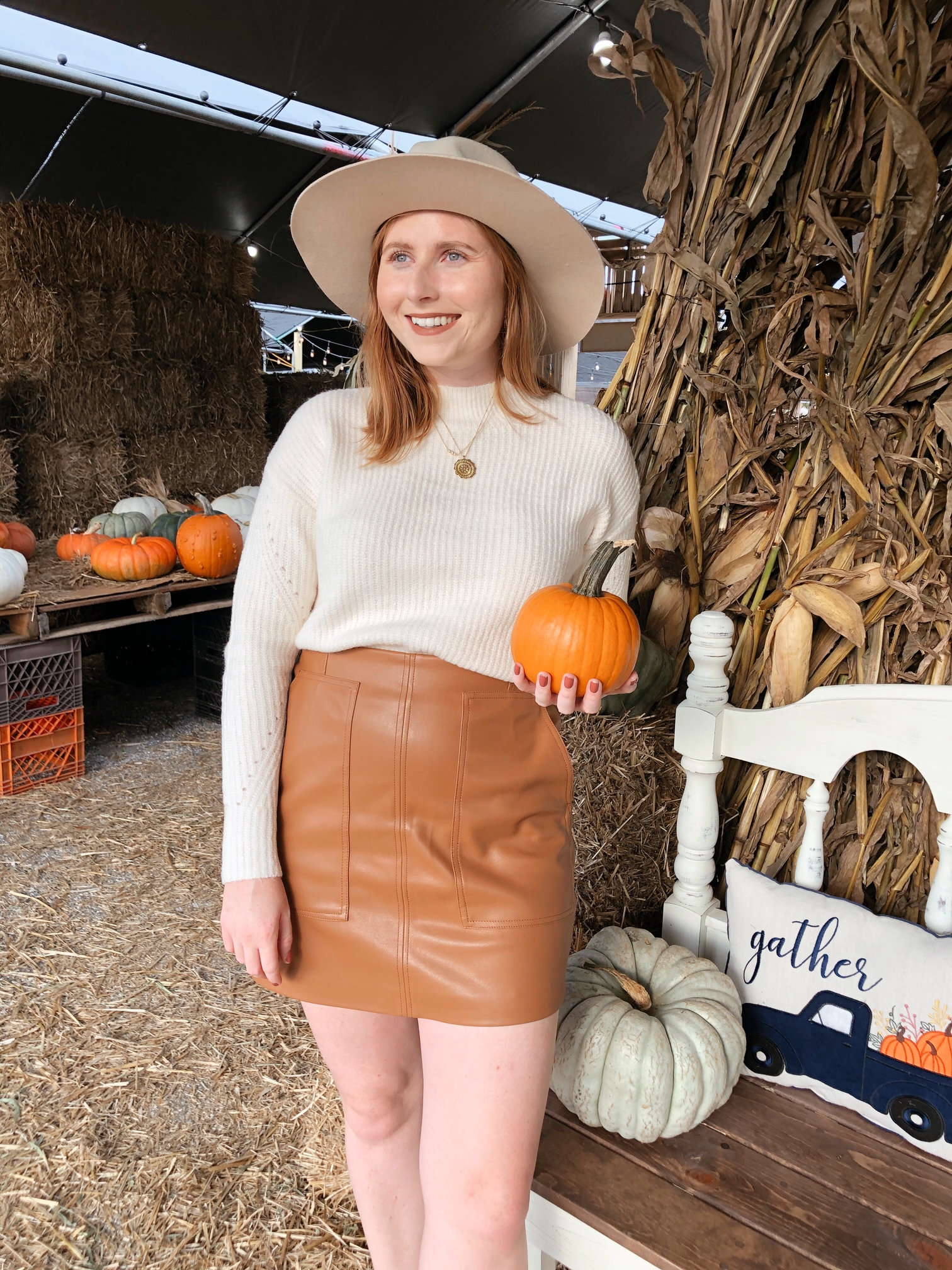 What To Wear to a Pumpkin Patch. Pumpkin Patch Outfit Ideas. Red haired woman wears a white knit long sleeve sweater tucked into a brown faux leather mini skirt. She is wearing brown suede lace-up ankle booties. She has on a wide-brim hat and is holding a pumpkin in her hand. 