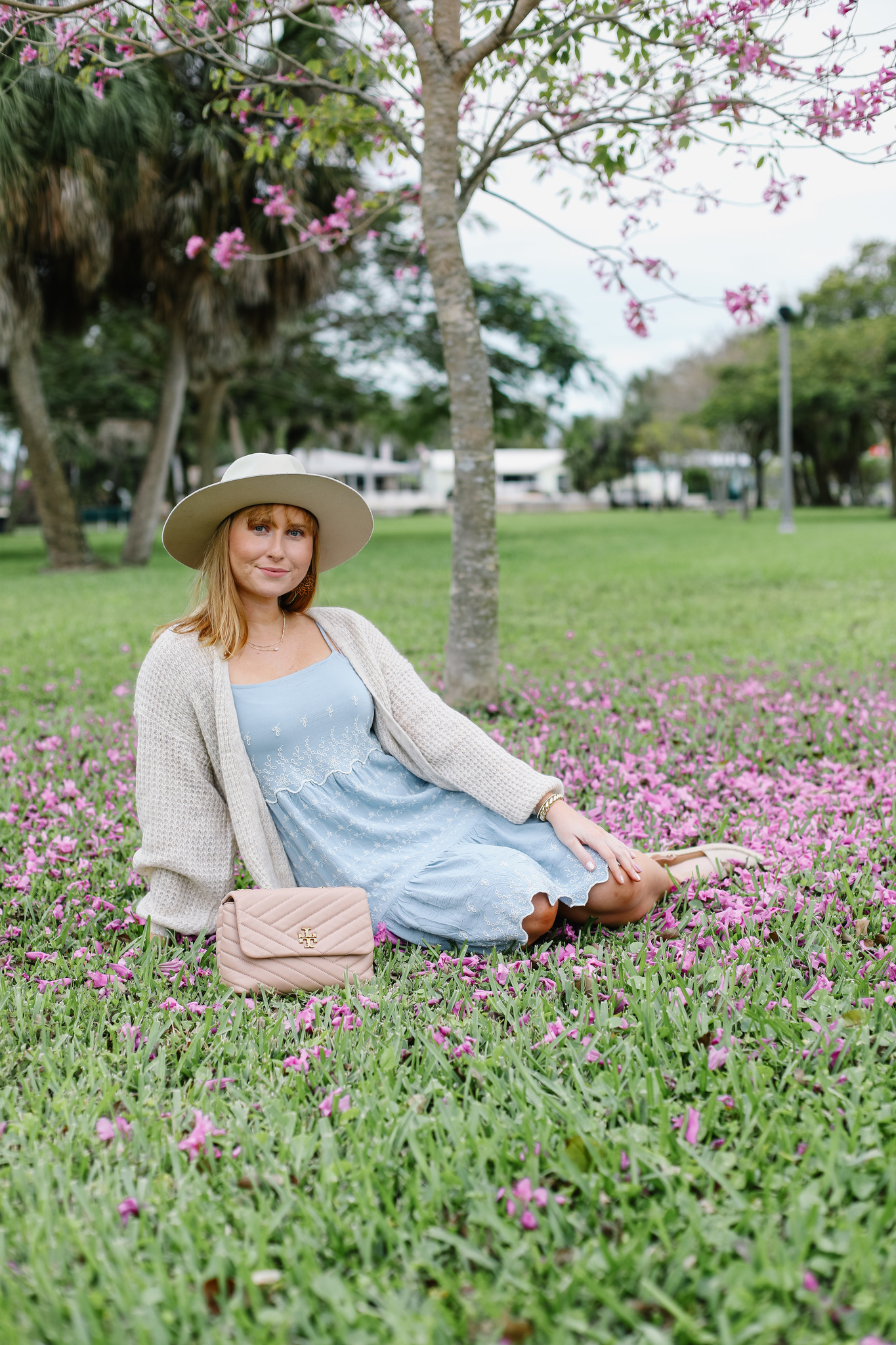 Amanda Burrows sits on grass with purple flowers. She is wearing a blue mini dress and light beige cardigan with a hat.