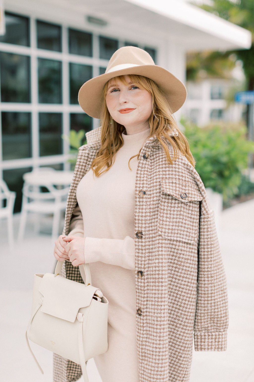 Affordable by Amanda wears a neutral sweater dress with a brown and cream houndstooth coat over her shoulders. She is holding a cream Celine nano belt bag and wearing a brown floppy felt hat on her head. 