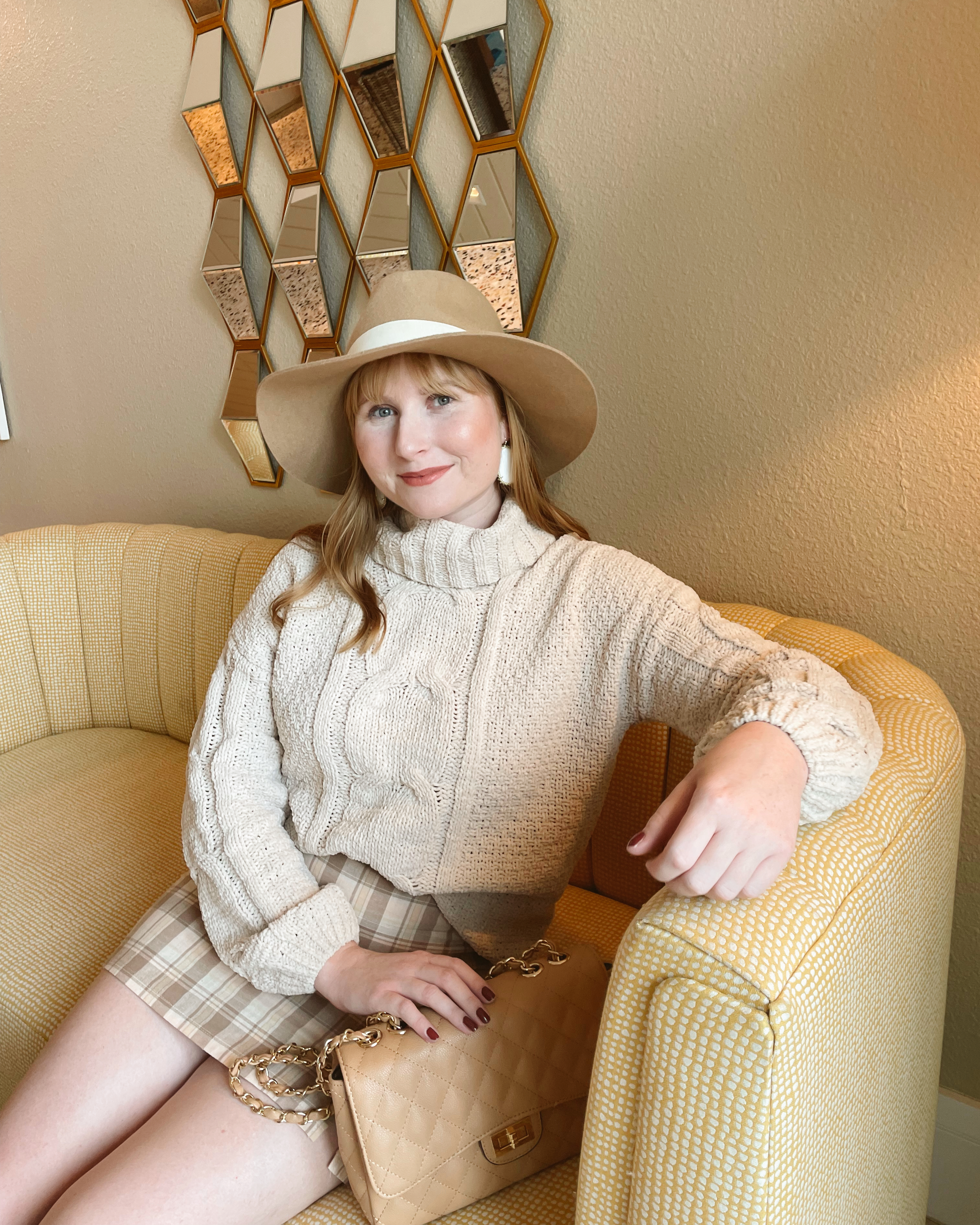 Affordable by Amanda wears a beige cream cable knit turtleneck sweater with a plaid mini skirt while sitting on a yellow couch